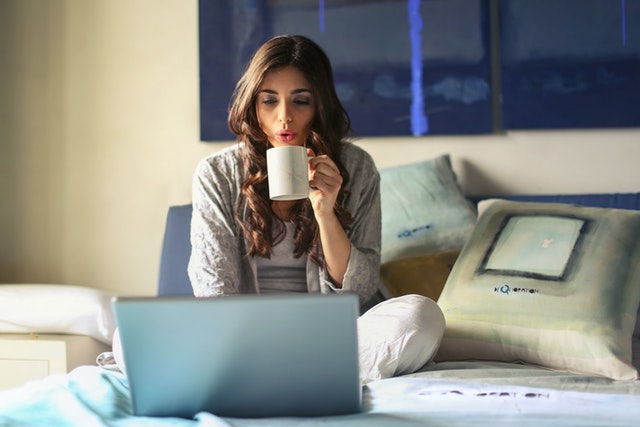 Women with a laptop sitting on her bed working and holding a coffee cup